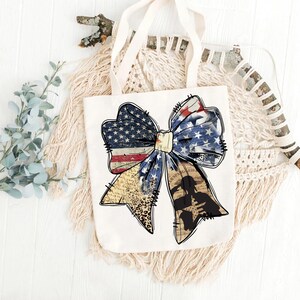 May include: A white tote bag with a large, distressed, red, white, and blue bow. The bow is made of fabric with a stars and stripes pattern. The bow is tied with a white ribbon. The bow is centered on the bag. The bag has a white background.
