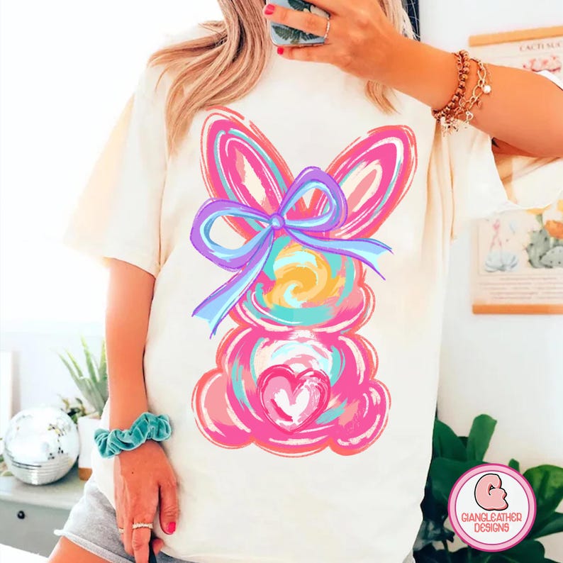 May include: A white t-shirt with a colorful abstract bunny design. The bunny is pink, blue, yellow, and green. It has a large bow on its head. The design is painted in a loose, whimsical style.