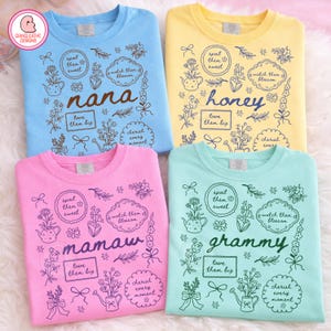 May include: Four crew neck sweatshirts in pastel colors: blue, yellow, pink, and mint green. Each shirt features a floral design with the words "nana", "honey", "mamaw", and "grammy" in a script font. The design includes the phrases "spoil them sweet", "watch them blossom", "love them big", and "cherish every moment".