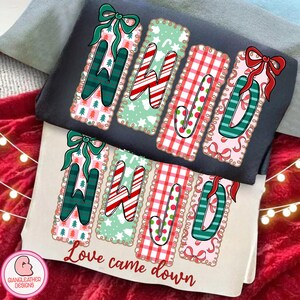 May include: Two t-shirts, one dark grey and one beige, are stacked. Each shirt features the word "WHOA" in a festive design with candy canes, bows, and Christmas trees. The text "Love came down" is printed on the beige shirt.