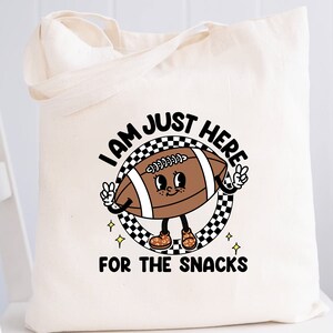 May include: A white canvas tote bag with a black and white checkered circle design. Inside the circle is a cartoon football with a face and arms. The football is wearing brown shoes with yellow laces. The text "I am just here for the snacks" is printed below the circle.