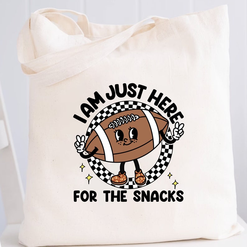 May include: A white canvas tote bag with a black and white checkered circle design. Inside the circle is a cartoon football with a face and arms. The football is wearing brown shoes with yellow laces. The text "I am just here for the snacks" is printed below the circle.