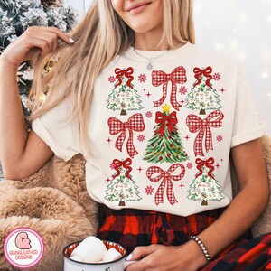 Puede incluir: Camiseta color crema con un dise&ntilde;o navide&ntilde;o. El dise&ntilde;o incluye &aacute;rboles de Navidad, lazos a cuadros rojos, copos de nieve y un &aacute;rbol de Navidad central adornado con una estrella y adornos. Parte de una colecci&oacute;n navide&ntilde;a.