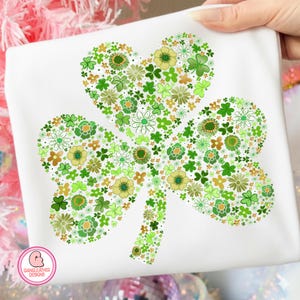 May include: White t-shirt featuring a large shamrock design made of various green and yellow flowers and clovers. The shamrock is centered on the shirt, with a heart-shaped top leaf. The design is held by a hand.