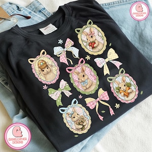 K&ouml;nnte beinhalten: Schwarzes T-Shirt mit ovalen Portr&auml;ts von Hasen, die Karotten, Blumen und Ostereier halten. Pastellfarbene Schleifen und florale Akzente umgeben die Portr&auml;ts. Das T-Shirt tr&auml;gt den Text "GIANGLEATHER DESIGNS".