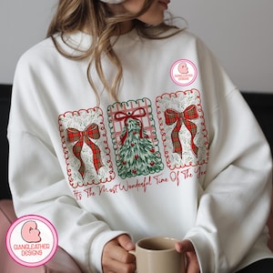May include: White sweatshirt featuring three Christmas-themed designs. The designs include a Christmas tree and plaid bows, framed with decorative borders. The text "It's The Most Wonderful Time Of The Year" is printed in red. The sweatshirt has the "Giangleather Designs" logo.