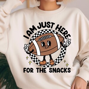 May include: A white sweatshirt with a cartoon football character wearing sneakers and the text "I am just here for the snacks".