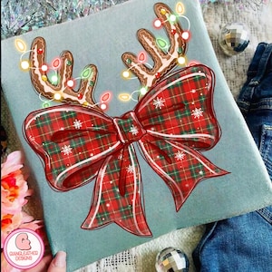 Puede incluir: Un coj&iacute;n decorativo azul claro con un dise&ntilde;o navide&ntilde;o. El dise&ntilde;o incluye cuernos de reno de pan de jengibre adornados con luces de cadena y un gran lazo a cuadros rojo con copos de nieve blancos. El coj&iacute;n tiene el texto "GIANGLEATHER DESIGNS".