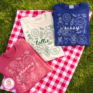 May include: Three sweatshirts in cream, rose, and navy blue are displayed on a red and white checkered picnic blanket. Each sweatshirt features floral designs and text, including "grammy," "lollie," and "sissy." The sweatshirts are on a green grassy background.