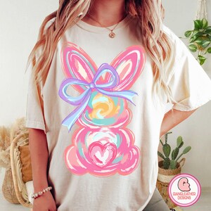 May include: A white t-shirt with a pink and blue bunny graphic. The bunny has a large bow and a heart on its chest.