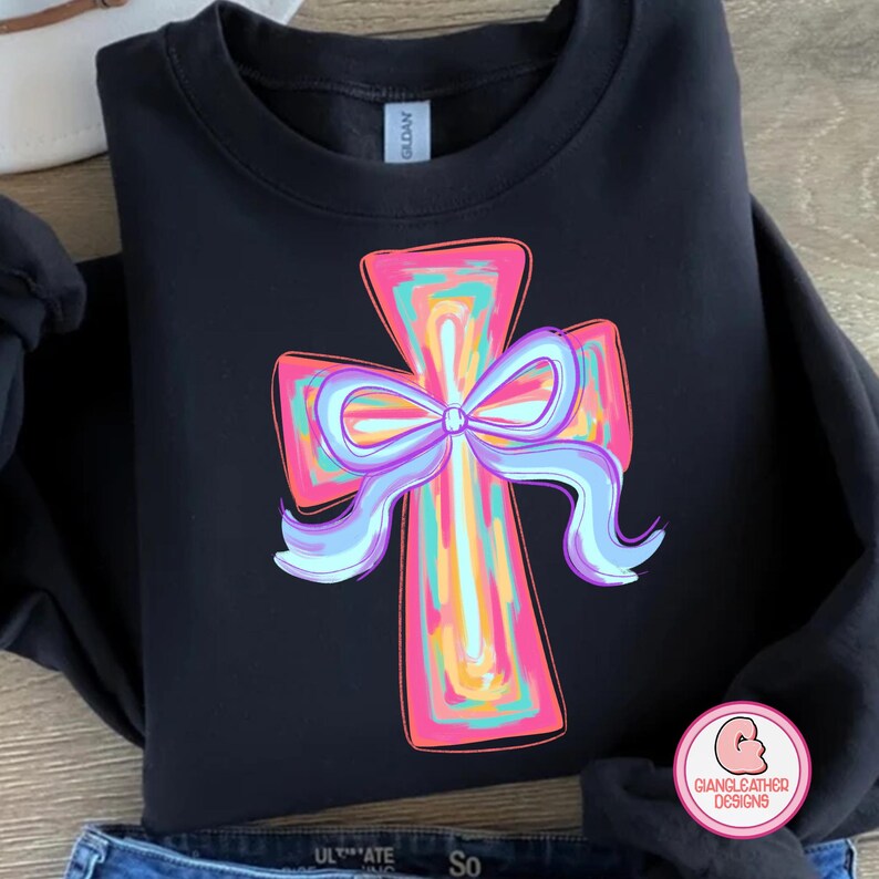 May include: Black sweatshirt with a colorful cross design featuring a purple bow. The cross is outlined in pink and has a rainbow-like pattern.