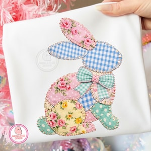 May include: A white fabric with a patchwork bunny design. The bunny features pink floral, blue gingham, and yellow floral patterns. A green and blue gingham bow adorns the bunny's neck. The design is stitched onto the fabric.