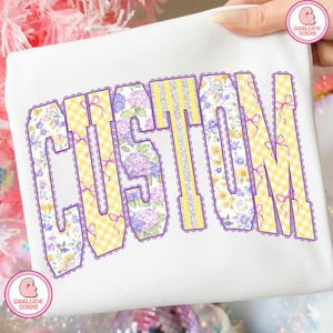 May include: White t-shirt with the word "CUSTOM" in a decorative font. The letters feature floral, gingham, and glitter patterns, outlined in purple. Small purple bows are also included in the design.
