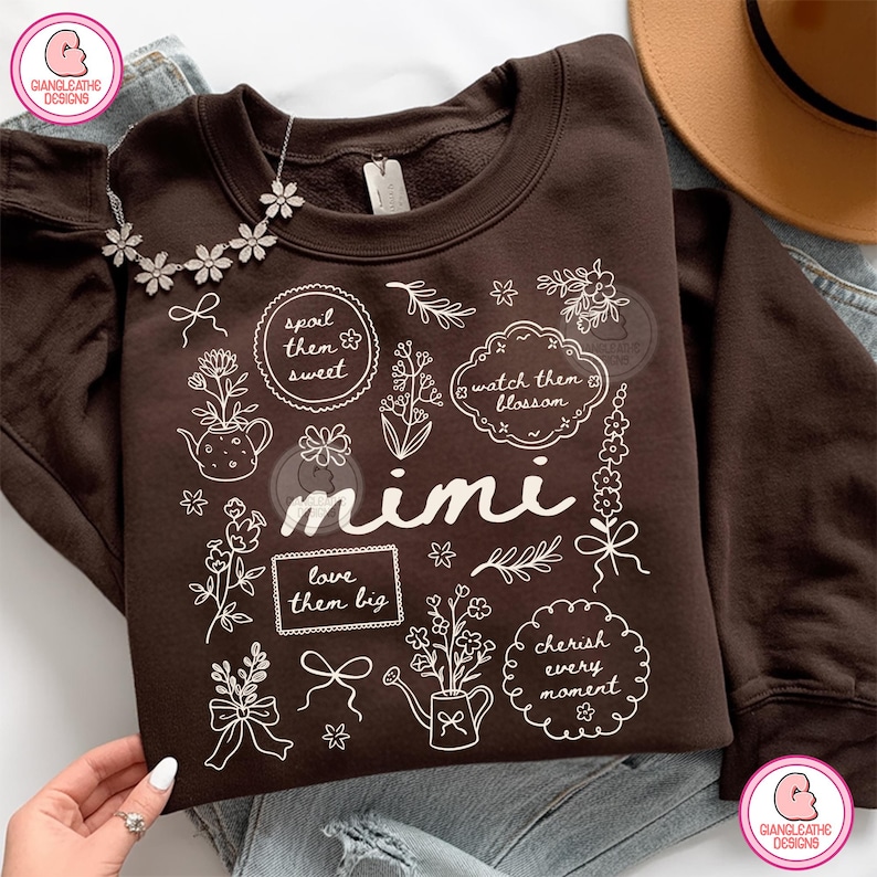 May include: A brown sweatshirt with white floral illustrations and the word "mimi" in a script font. The design includes phrases like "spoil them sweet" and "cherish every moment". A silver necklace with flower pendants is draped on the sweatshirt.
