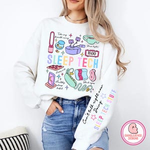 Könnte beinhalten: Weißer Pullover mit einem bunten Grafikdesign mit Schlaftechnologie-Artikeln wie einer Augenmaske, Schlafsounds und einem Schlaf-Tracker. Der Text "SLEEP TECH" ist in Regenbogenfarben gedruckt. Der Ärmel trägt den Text "Can't talk right now Doing SLEEP TECH STUFF".