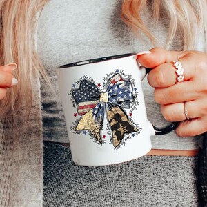 May include: White ceramic mug with a black rim and handle. The mug features a red, white, and blue bow with a silhouette of a soldier in the center. The bow is surrounded by the text "Honor the brave" repeated multiple times.