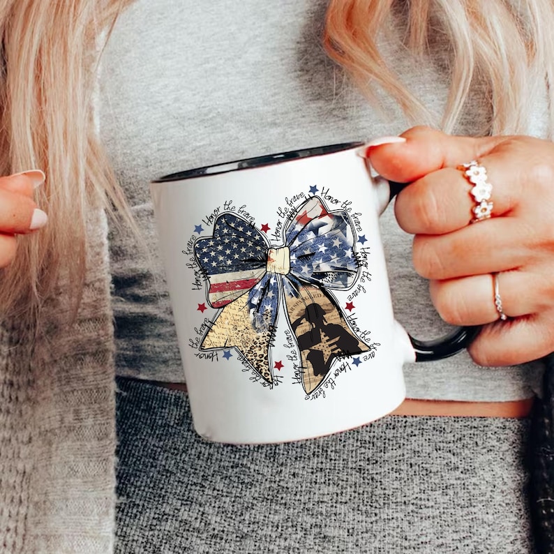 May include: White ceramic mug with a black rim and handle. The mug features a red, white, and blue bow with a silhouette of a soldier in the center. The bow is surrounded by the text "Honor the brave" repeated multiple times.