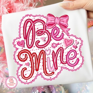 Puede incluir: Camiseta blanca con un diseño de San Valentín. El diseño presenta las palabras "Be Mine" en escritura roja y rosa, rodeadas de corazones y un lazo rosa. El diseño está enmarcado por un borde festoneado.