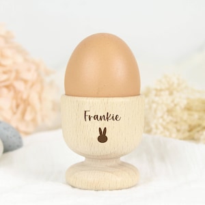 May include: A wooden egg cup with a bunny design and the name Frankie engraved on it. The egg cup is holding a brown egg.