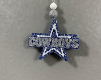 May include: A blue and silver glitter star-shaped ornament with the word "COWBOYS" written on it. The ornament is hanging from a white string with silver and white beads.