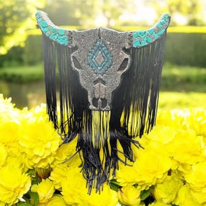 May include: A decorative bull skull with turquoise horns and a central turquoise and silver design. The skull is embellished with silver accents and black fringe, set against a backdrop of yellow flowers and greenery.