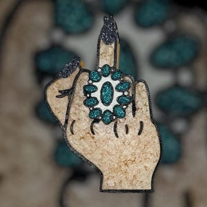 May include: A hand-shaped pin with a raised middle finger. The pin features a beige textured surface, black outlines, and glitter accents on the fingernails and a ring. The ring has a white center and teal glitter stones.