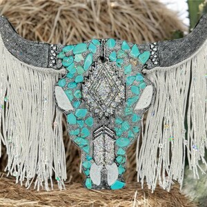 May include: A turquoise and silver embellished bull skull with white fringe. The skull is decorated with turquoise stones and rhinestones.