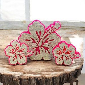 May include: Three glittery hibiscus flower decorations. The flowers have a gold glitter center and petals, with a bright pink outline. The flowers are arranged on a rustic wooden surface, with a window in the background.