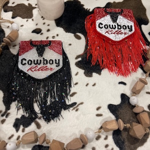 May include: Two red and white beaded cowboy killer patches with black and red fringe. The patches are on a white and brown cowhide background.
