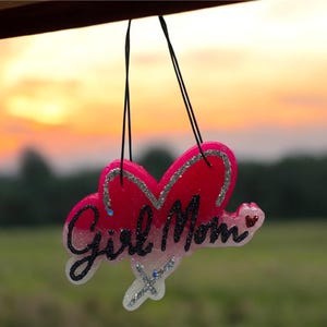 May include: A heart-shaped ornament with the words "Girl Mom" in cursive. The ornament is primarily pink with silver glitter accents and hangs from black strings against a sunset backdrop.