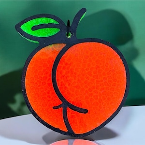 May include: A black and orange peach shaped ornament with a green leaf. The peach is outlined in black and has a textured surface.