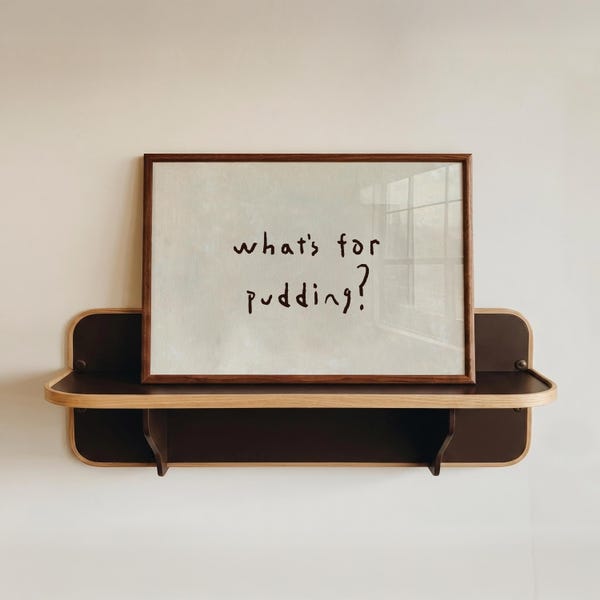 以下が含まれることがあります： 明るいベージュの背景に「what's for pudding?」というダークブラウンの手書き文字が中央に配置された、長方形の額入りプリント。フレームはダークウッド製で、角が丸いダークブラウンの壁掛け棚の上に置かれています。