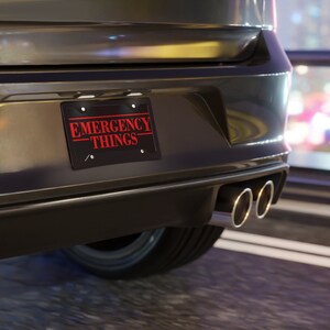 Peut inclure: Plaque d'immatriculation noire avec les mots "EMERGENCY THINGS" en rouge, montée à l'arrière d'une voiture gris foncé. La plaque a un fond noir avec des lettres rouges. La voiture a des tuyaux d'échappement doubles.