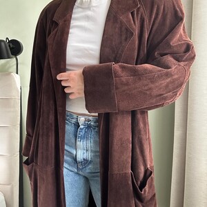 May include: A long brown suede coat with a collar and pockets. The coat is worn over a white t-shirt and blue jeans.