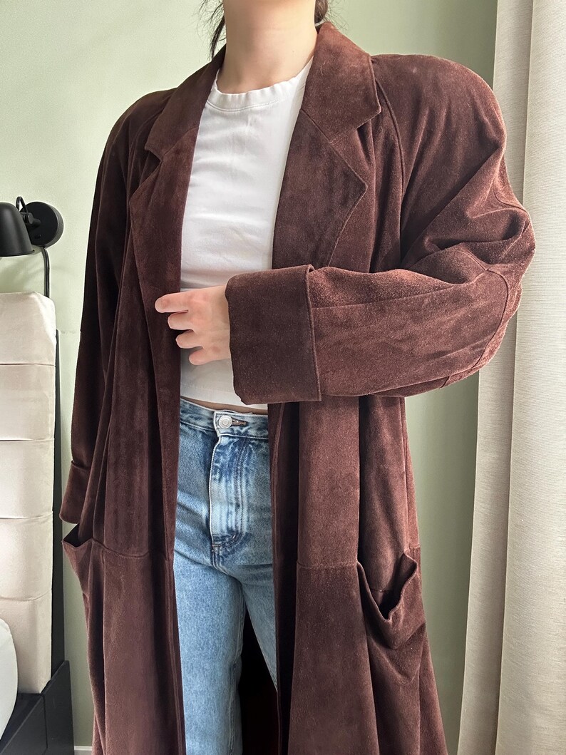 May include: A long brown suede coat with a collar and pockets. The coat is worn over a white t-shirt and blue jeans.