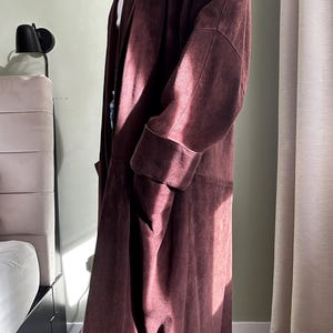 May include: A long, burgundy suede coat with a shawl collar and long sleeves. The coat is unbuttoned and has a relaxed fit.