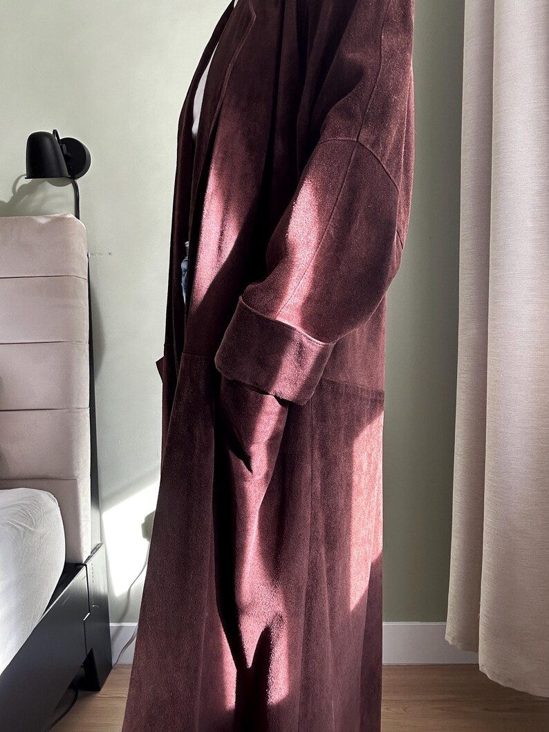 May include: A long, burgundy suede coat with a shawl collar and long sleeves. The coat is unbuttoned and has a relaxed fit.
