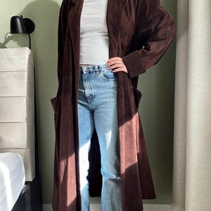 May include: A long brown suede coat with a collar and pockets. The coat is worn over a white t-shirt and blue jeans.