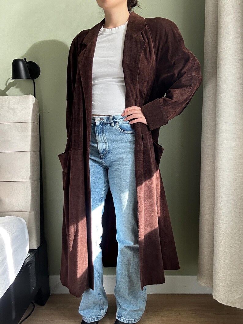 May include: A long brown suede coat with a collar and pockets. The coat is worn over a white t-shirt and blue jeans.