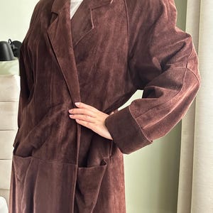 May include: A long brown suede coat with a collar and pockets. The coat has a relaxed fit and a long length.