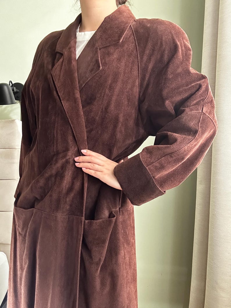 May include: A long brown suede coat with a collar and pockets. The coat has a relaxed fit and a long length.