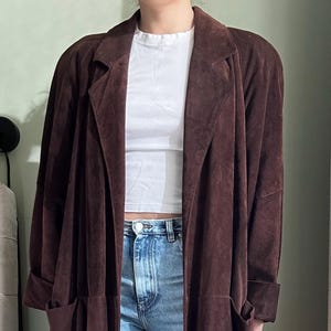May include: A long brown suede jacket with a collar and pockets. The jacket is open and the person wearing it is wearing a white t-shirt and blue jeans.