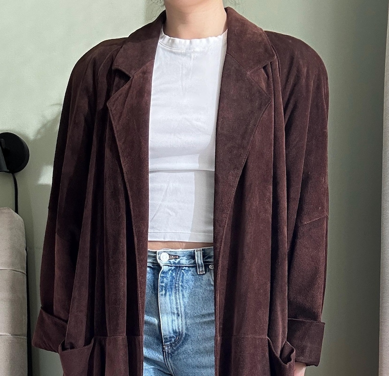 May include: A long brown suede jacket with a collar and pockets. The jacket is open and the person wearing it is wearing a white t-shirt and blue jeans.