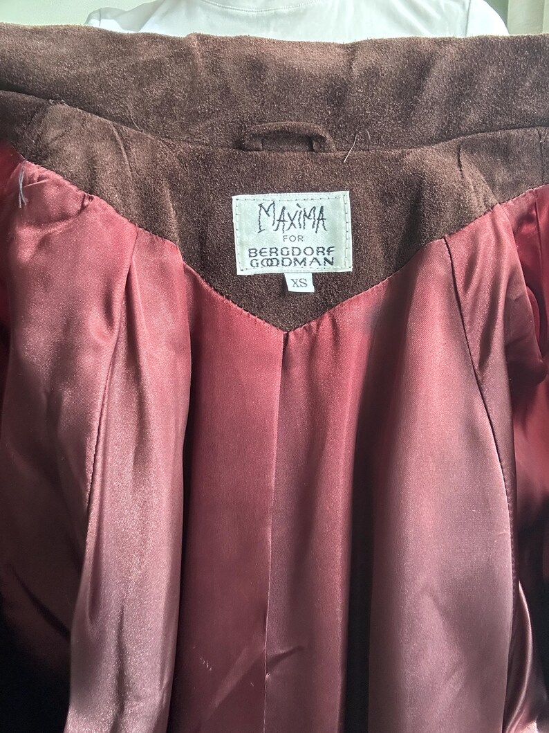May include: A brown suede jacket with a burgundy lining. The jacket has a label that reads "Maxima for Bergdorf Goodman" and "XS".
