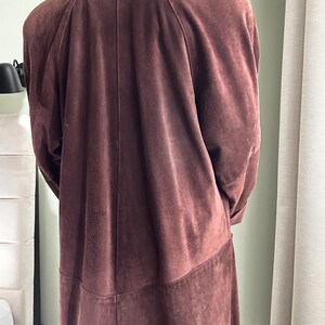 May include: A long brown suede coat with a classic collar and a straight cut. The coat has a smooth texture and a slightly textured finish.