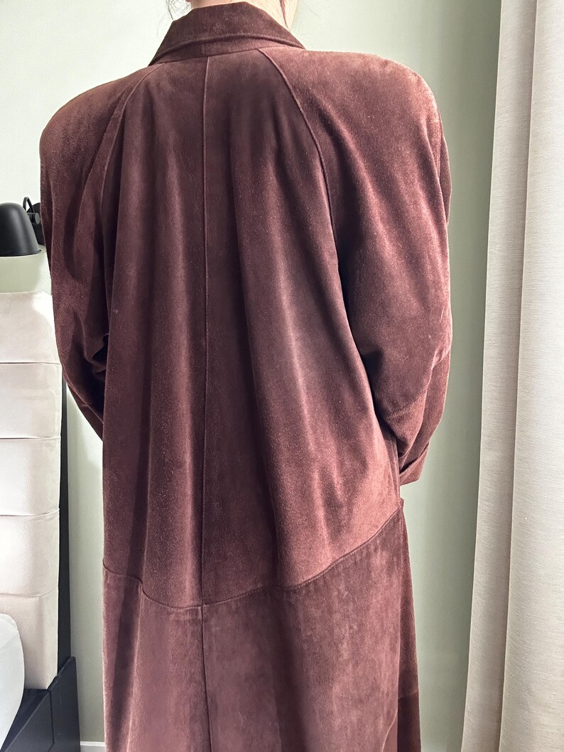 May include: A long brown suede coat with a classic collar and a straight cut. The coat has a smooth texture and a slightly textured finish.