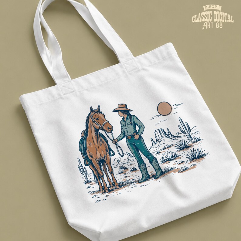 May include: White tote bag featuring a desert scene with a cowgirl and horse. The illustration includes cacti, mountains, and a sun. The bag has the text "SHOP CLASSIC DIGITAL ART 88" in the upper right corner.