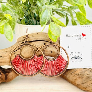 May include: A pair of handmade earrings with red and white fringe hanging from a copper-toned hoop. The earrings are displayed on a wooden surface with a card that reads "Handmade with love" and "Emily's Jams".