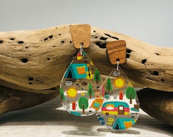 May include: A pair of teardrop-shaped earrings with a wood backing. The earrings are clear and feature a colorful camping-themed design with a camper, tent, trees, and a sun.