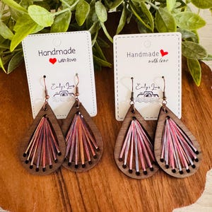 May include: Wooden teardrop earrings with colorful thread accents. Each earring features a dark wood base with pink, orange, and purple thread radiating from the center. The earrings are displayed on a wooden surface with a tag that reads "Handmade with love."